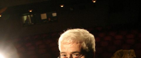 Valkohiuksinen ja silmälasipäinen henkilö näkyy osittain hämärässä teatterin etualalla, jonka taustalla on tyhjiä punaisia istuimia ja ylävaloja.