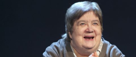 Vanhempi nainen, jolla on lyhyet harmaat hiukset, hymyilee lämpimästi katsoen hieman ylöspäin tummaa taustaa vasten. Hänellä on yllään vaalea pusero ja ruskea neuletakki.