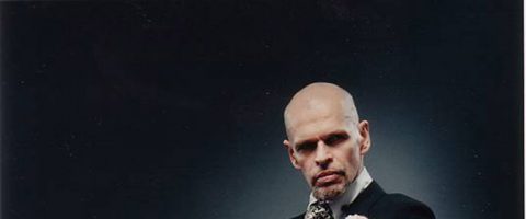 Mustaan pukuun ja kuviolliseen solmioon pukeutunut kalju mies katsoo vakavana kameraan tummaa taustaa vasten.