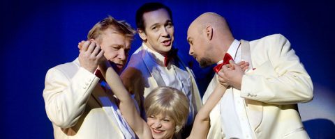 Neljä valkoisiin pukuihin ja punaisiin rusetteihin pukeutunutta ihmistä poseeraa dramaattisesti lavalla; kolme miestä pitelee hymyilevän lyhythiuksisen, vaalean naisen käsiä kirkkaan sinisessä valaistuksessa.