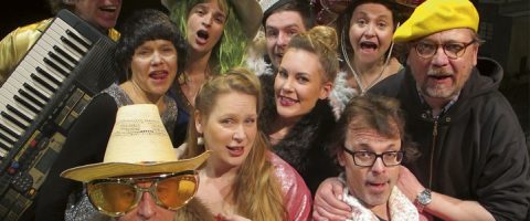 Kymmenen aikuisen ryhmä, jolla on värikkäitä ja omituisia pukuja, kuten hattuja, peruukkeja, aurinkolaseja ja höyhenboa, poseeraa tiiviisti yhdessä ja hymyilee kameralle. Yhdellä henkilöllä on kädessään näppäimistö.