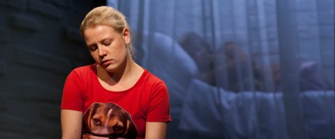 Vaaleatukkainen nainen, jolla on punainen paita, jossa on koiran kasvot, istuu silmät kiinni ja katsoo alaspäin. Taustalla, läpikuultavien verhojen takana, joku makaa sängyssä, osittain peitettynä.