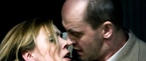 Lähikuva kahdesta ihmisestä, naisesta ja miehestä, joilla on voimakkaat kasvonilmeet ja jotka nojaavat tiiviisti toisiaan kohti, mikä viittaa jännitteeseen tai yhteenottoon. Tausta on tumma ja epätarkka.