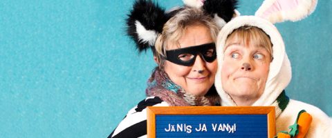 Kaksi vanhempaa naista puvuissa poseeraa yhdessä. Toisella on mustavalkoiset eläinkorvat ja musta naamio, toisella valkoinen pupupuku, jossa on porkkana kädessä. He hymyilevät ja pitelevät kylttiä, jossa lukee JANIS JA VANNI.