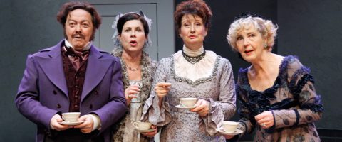 Neljä viktoriaaniseen tyyliin pukeutunutta ihmistä, joilla on kädessään teekupit ja lautaset, seisovat lähekkäin ja katsovat hämmästyneinä eteenpäin, mikä viittaa teatteriesityksen kohtaukseen.