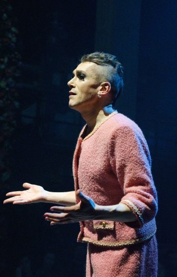 Vaaleanpunaiseen kuvioituun asuun pukeutunut henkilö tekee eleitä avoimin käsin ja ilmeikkäin kasvonpiirtein seisoen dramaattisen näyttämövalaistuksen alla pimeässä teatteritilassa.