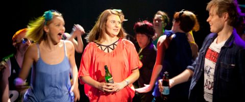 Punatoppinen nainen seisoo juoma kädessään ja näyttää kömpelöltä, kun ihmiset hänen ympärillään tanssivat innokkaasti ja ovat vuorovaikutuksessa vilkkaassa, värikkäässä juhlakuvassa.