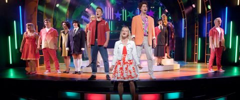 Ryhmä värikkäisiin pukuihin pukeutuneita esiintyjiä seisoo ja laulaa kirkkaasti valaistulla näyttämöllä, jossa on neonvalot ja elävä tausta, kun taas kukkamekkoon pukeutunut nainen seisoo edessä, keskellä näyttämöä.