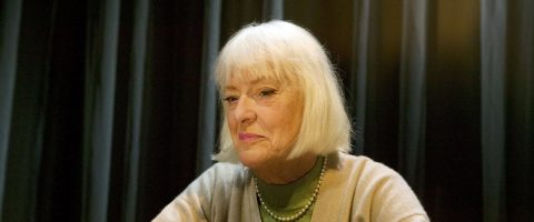 Vanhempi nainen, jolla on lyhyet valkoiset hiukset ja beige pusero, istuu tummien, pehmeästi valaistujen verhojen edessä, katsoo hieman sivulle ja hymyilee lempeästi.