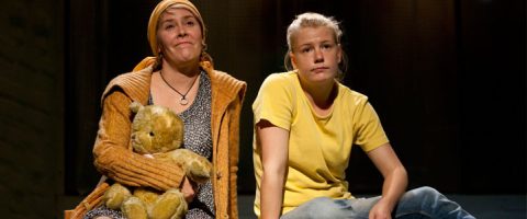 Kaksi naista istuu vierekkäin. Toisella on päähuivi, ja hänellä on nalle kädessään. Hän on pukeutunut kuviomekkoon ja oranssiin puseroon. Toisella on keltainen t-paita ja hän katsoo neutraalilla ilmeellä eteenpäin. Molemmat vaikuttavat mietteliäiltä.