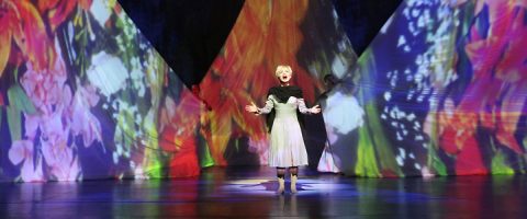 Esiintyjä seisoo näyttämön keskellä, dramaattisesti valaistuna, yllään valkoinen mekko ja tumma huivi, ja heidän takanaan on elinvoimaisia, värikkäitä kukkakuvioita, jotka on heijastettu suurille kolmionmuotoisille taustakuvioille.