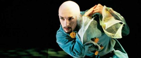 Siniseen pukuun pukeutunut esiintyjä, jolla on läpikuultavat hihat ja keltaiset korostukset, poseeraa dramaattisesti kädet yhdessä ja katsoo intensiivisesti eteenpäin. Tausta on tumma, mikä korostaa teatterillista ilmettä.