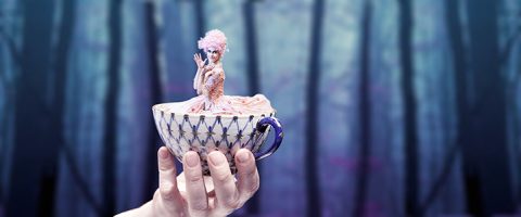 Kädessä on kuvioitu teekuppi, jossa on pieni nainen, joka on pukeutunut hienostuneeseen vaaleanpunaiseen pukuun ja peruukkiin, ja taustalla on unenomainen, sumea metsä.