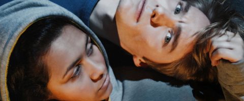 Kaksi ihmistä makaa lähekkäin maassa, päät koskettavat toisiaan. Toisella on harmaa huppari, toisella on vaaleat hiukset ja hän katsoo ylöspäin. Molemmilla on rauhalliset, mietteliäät ilmeet pehmeässä valaistuksessa.