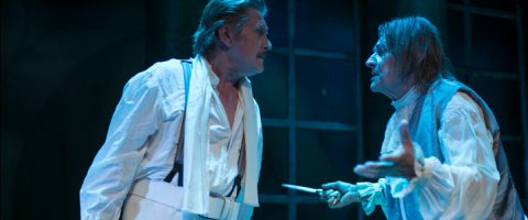 Kaksi historiallisiin pukuihin pukeutunutta näyttelijää käyvät jännittävää keskustelua näyttämöllä; toinen elehtii käsillään pitäen kädessään veistä, ja molemmilla on vakavat ilmeet dramaattisessa sinisessä valaistuksessa.