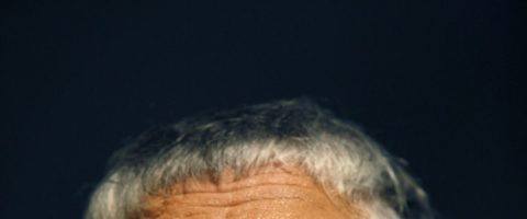 Lähikuva, jossa näkyy henkilön pään yläosa, jossa on lyhyet, harmaat hiukset tummaa taustaa vasten. Vain otsa ja hiukset ovat näkyvissä.