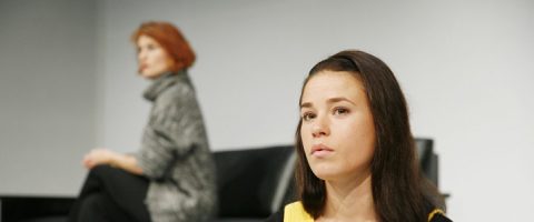 Kaksi naista istuu erillään sisätiloissa; etualalla olevalla naisella on pitkät tummat hiukset ja hän katsoo vakavana eteenpäin, kun taas taustalla olevalla, hieman epäselvästi näkyvällä naisella on lyhyet punaiset hiukset ja hänellä on harmaa villapaita.