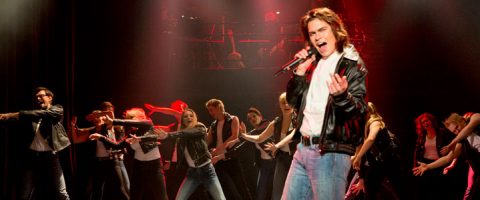 Ryhmä esiintyjiä tanssii energisesti lavalla punaisessa valaistuksessa, ja edessä on nahkatakkinen henkilö, joka laulaa intohimoisesti mikrofoniin.