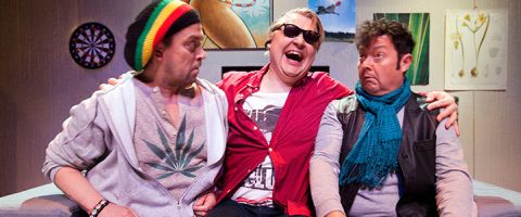 Kolme miestä istuu lähekkäin sohvalla, nauravat ja tekevät yllättyneitä ilmeitä. Vasemmalla olevalla miehellä on pipo ja huppari, jossa on kannabiksen lehti, keskimmäisellä miehellä on aurinkolasit ja punainen paita, ja oikealla miehellä on sininen huivi.