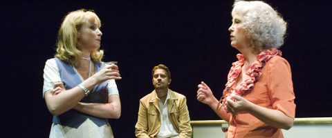 Lavalla on kolme aikuista. Etualalla kaksi naista keskustelee, toisella on muki kädessään. Taustalla istuu mies, joka tarkkailee heitä. Kohtaus on dramaattinen ja näyttää olevan osa teatteriesitystä.