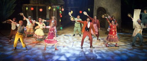 Ryhmä värikkäisiin pukuihin pukeutuneita näyttelijöitä esiintyy energisesti lavalla, hattuja ilmassa, ja taustalla on juhlavaloja ja puisia lavasteita. Kohtaus vaikuttaa eloisalta ja teatraaliselta.