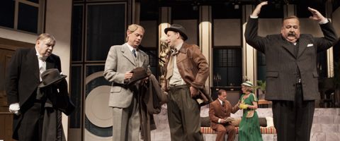 Neljä vintage-vaatteisiin pukeutunutta miestä seisoo näyttämön edessä, ja yksi heistä nostaa dramaattisesti kätensä ylös. Kaksi vihreisiin mekkoihin pukeutunutta naista istuu sohvalla taustalla. Kohtaus näyttää olevan menneisyyteen sijoittuvasta teatterinäytelmästä.