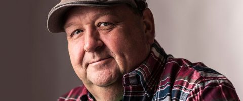 Vanhempi mies, jolla on harmaa lippis ja punainen ruutupaita, hymyilee lempeästi kameralle pehmeästi valaistua, neutraalia taustaa vasten.