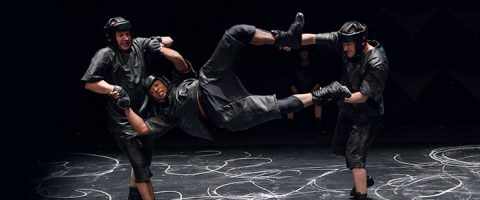 Kolme mustiin suojavarusteisiin pukeutunutta henkilöä tekee dynaamisen akrobaattisen liikkeen; kaksi pitää kolmatta henkilöä vaakatasossa maanpinnan yläpuolella lavalla, jossa on liitumaisia merkintöjä.