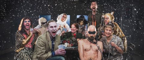 Joukko värikkäisiin teatteripukuihin pukeutuneita ihmisiä nauraa ja reagoi dramaattisesti, kun heidän ympärilleen putoaa tekolunta; yksi henkilö on ilman paitaa ja silmänaamari, ja toinen on pukeutunut muumioksi.