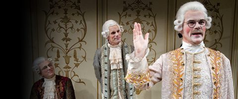 Kolme koristeltuihin 1700-luvun pukuihin ja valkoisiin peruukkeihin pukeutunutta miestä seisoo kuvioitua seinää vasten; yksi edessä seisovista miehistä tekee kädellään eleitä, kun kaksi muuta katselee.