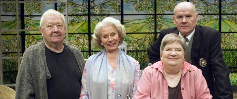 Neljä iäkästä aikuista, kolme istumassa ja yksi takana seisomassa, poseeraavat sisätiloissa värikkään lasimaalauksen ja luontokuvan edessä. Kaikki hymyilevät ja ovat pukeutuneet villapaitoihin tai juhlavaatteisiin.