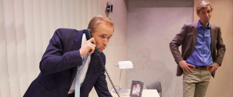 Pukumies nojaa työpöydän yli ja puhuu keskittyneenä langalliseen puhelimeen. Toinen mies seisoo taustalla kädet lanteilla ja tarkkailee häntä. Toimistossa on lamppu, kehystetty valokuva ja pystysuuntaiset kaihtimet.