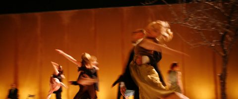 Ryhmä tanssijoita esiintyy lavalla sumealla liikkeellä luoden dynaamisen ja energisen kohtauksen lämpimällä, kultaisella taustalla. Kuvan oikealla puolella näkyy lehdetön puu.