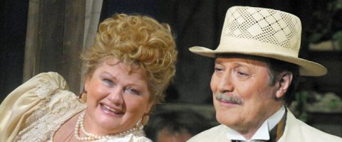 Nainen, jolla on kiharat vaaleat hiukset ja helmikaulakoru, hymyilee viiksekkään miehen vieressä, jolla on vaalea hattu ja puku. Molemmat näyttävät pukeutuneen vintage- tai teatteripukuihin.