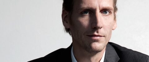 Tummaraitaiseen pukuun ja vaaleaan paitaan pukeutunut mies katsoo kameraan. Hänellä on vaalea iho, siniset silmät ja lyhyet ruskeat hiukset, ja hän on poseerannut vaaleaa taustaa vasten.