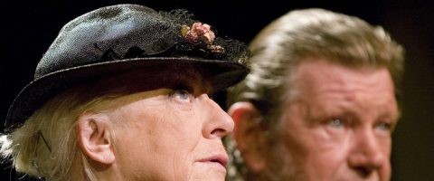 Iäkäs nainen, jolla on musta hattu, jossa on pieni kukkakoriste, katsoo mietteliäästi eteenpäin, kun taas taustalla näkyy epäselvästi iäkäs parrakas mies.