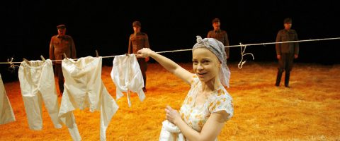 Huivipäinen nainen ripustaa valkoisia vaatteita narulle hymyillen, ja taustalla seisoo neljä univormupukuista miestä oljilla päällystetyllä lavalla.