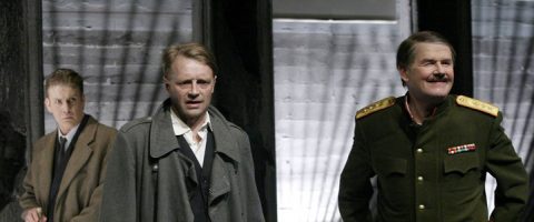 Lavalla seisoo kolme miestä; yhdellä on puku ja päällystakki, toisella pitkä harmaa takki ja kolmannella koristeltu sotilasunivormu ja viikset. Taustalla on kuvioitu harmaa seinä.