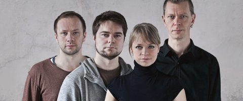 Neljä aikuista seisoo tiiviisti yhdessä vaaleaa, kuvioitua seinää vasten. Kolme miestä ja yksi nainen katsovat suoraan kameraan neutraalilla ilmeellä. Nainen seisoo edessä, yllään musta poolokaulus.