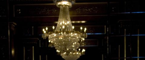 Suuri kristallikruunu loistaa lämpimästi tummassa, koristeellisesti sisustetussa huoneessa, jossa on tummaa puupanelointia. Kohtaus on hämärästi valaistu, mikä korostaa kattokruunun kimaltelevaa lasia ja monimutkaista muotoilua.