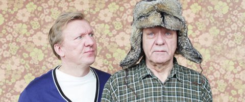 Kaksi miestä seisoo kukkakuvioisen seinän edessä; vasemmalla oleva mies katsoo oikealla olevaa miestä, jolla on karvainen talvihattu ja ruutupaita, tuijottaen tyhjin silmin eteenpäin.