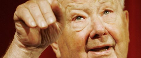 Iäkäs mies, jolla on vaalea iho ja lyhyet valkoiset hiukset, tekee eleitä oikealla kädellään, sormet taivutettuina, ja katsoo hymyillen ylöspäin. Tausta on punainen ja epätarkka.