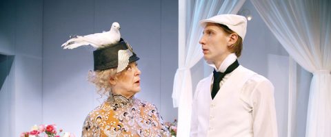 Vanhempi nainen, jolla on musta hattu, jonka päällä on valkoinen lintu, katsoo valkoiseen juhla-asuun ja lippikseen pukeutunutta miestä; molemmat seisovat valkoisten verhojen ja kukkien edessä ja vaikuttavat yllättyneiltä tai hämmentyneiltä.