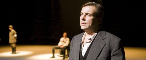 Pukumies seisoo etualalla valokeilan alla ja näyttää mietteliäältä. Taustalla näkyy kaksi muuta ihmistä hämärästi valaistulla näyttämöllä, toinen seisoo ja toinen istuu.