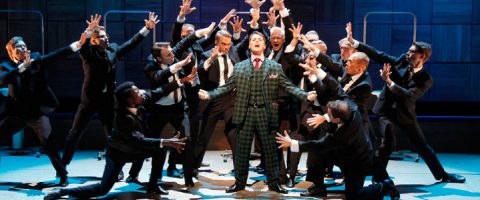 Ruudulliseen pukuun pukeutunut mies seisoo näyttämön keskellä kädet ojennettuina, ja häntä ympäröi joukko tummiin pukuihin pukeutuneita miehiä, jotka kurottautuvat dramaattisesti häntä kohti teatteriesityksessä.