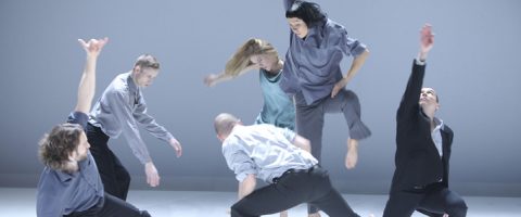 Kuusi tanssijaa väljissä, neutraalisävyisissä vaatteissa tekee ilmeikkäitä, dynaamisia liikkeitä minimalistisella, vaalealla näyttämöllä, jonka tausta on yksivärinen. Jotkut kurottautuvat ylöspäin, kun taas toiset kumartuvat tai nojaavat, mikä luo liikkeen ja energian tunnetta.