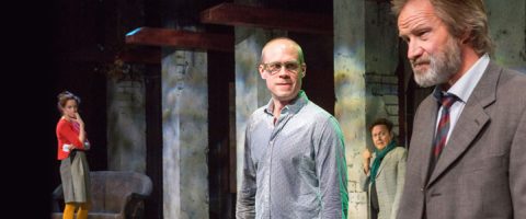 Neljä ihmistä on näyttämöllä dramaattisessa kohtauksessa. Yksi pukumies seisoo etualalla ja näyttää vakavalta; hänen takanaan on silmälasipäinen mies, joka katsoo eteenpäin. Kaksi muuta seisoo kauempana takana, maalaispylväiden lähellä, ja tarkkailee kohtausta.