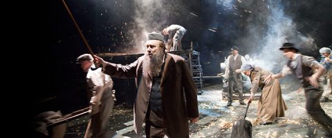 Parrakas mies aikakauden puvussa elehtii kepillä savuisella näyttämöllä, jota ympäröivät 1900-luvun alun asuihin pukeutuneet näyttelijät esittävät energisen kohtauksen rekvisiitan ja dramaattisen valaistuksen avulla.