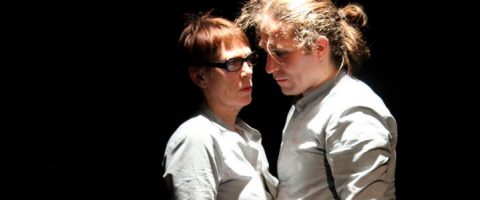 Kaksi ihmistä seisoo lähekkäin dramaattisessa valaistuksessa, molemmilla on vaaleat paidat. Toisella henkilöllä on lyhyet hiukset ja silmälasit, kun taas toisella on pitkät hiukset taakse sidottuina. He näyttävät jakavan intensiivisen hetken.