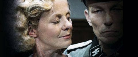 Vaaleatukkainen nainen, jolla on lempeä hymy, seisoo lähellä SS-sotilaspukuun pukeutunutta ankarakasvoista miestä, ja molemmat katsovat hieman toisiaan kohti tummaa taustaa vasten.